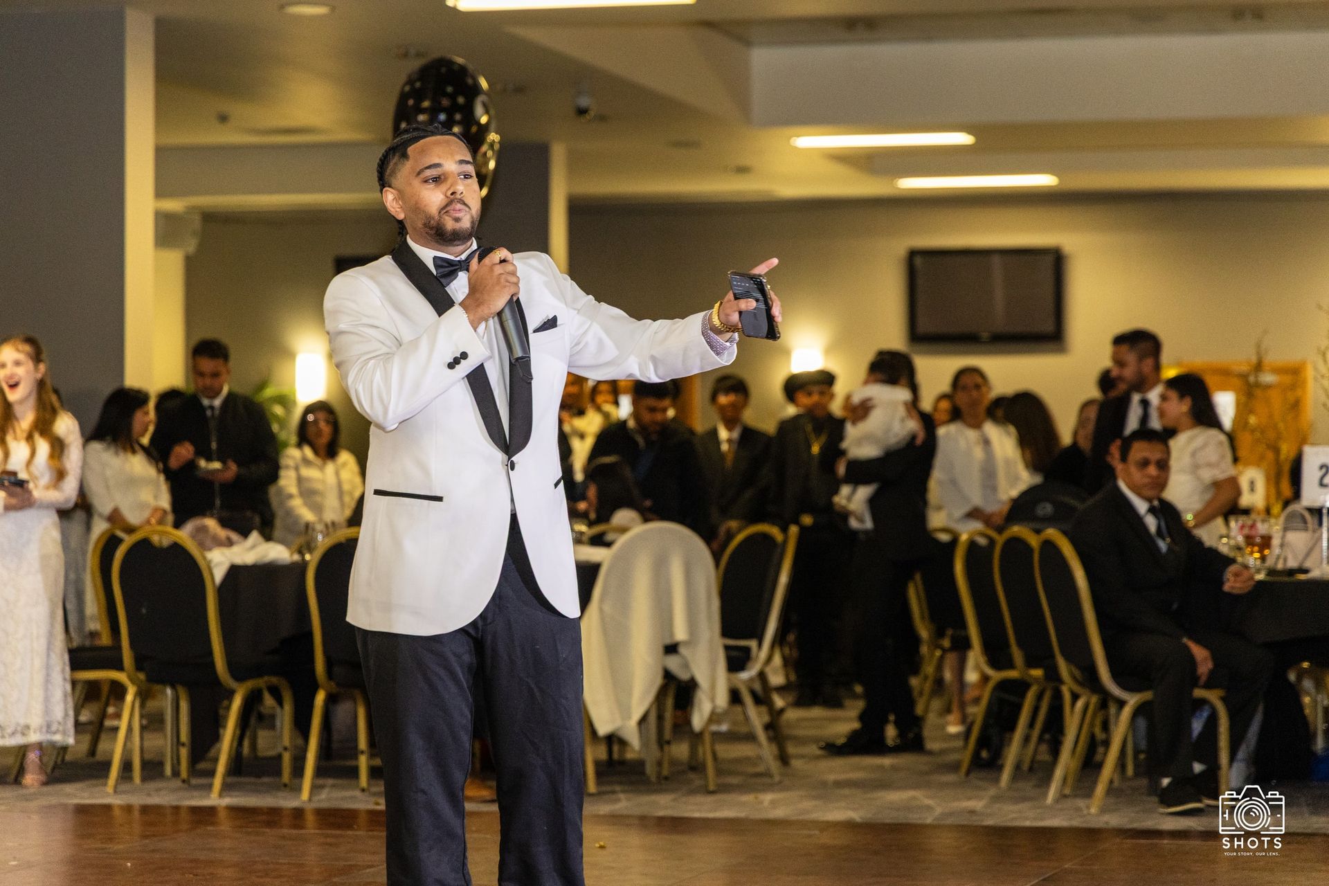 A man in a white dinner jacket holding a microphone at a seated celebration