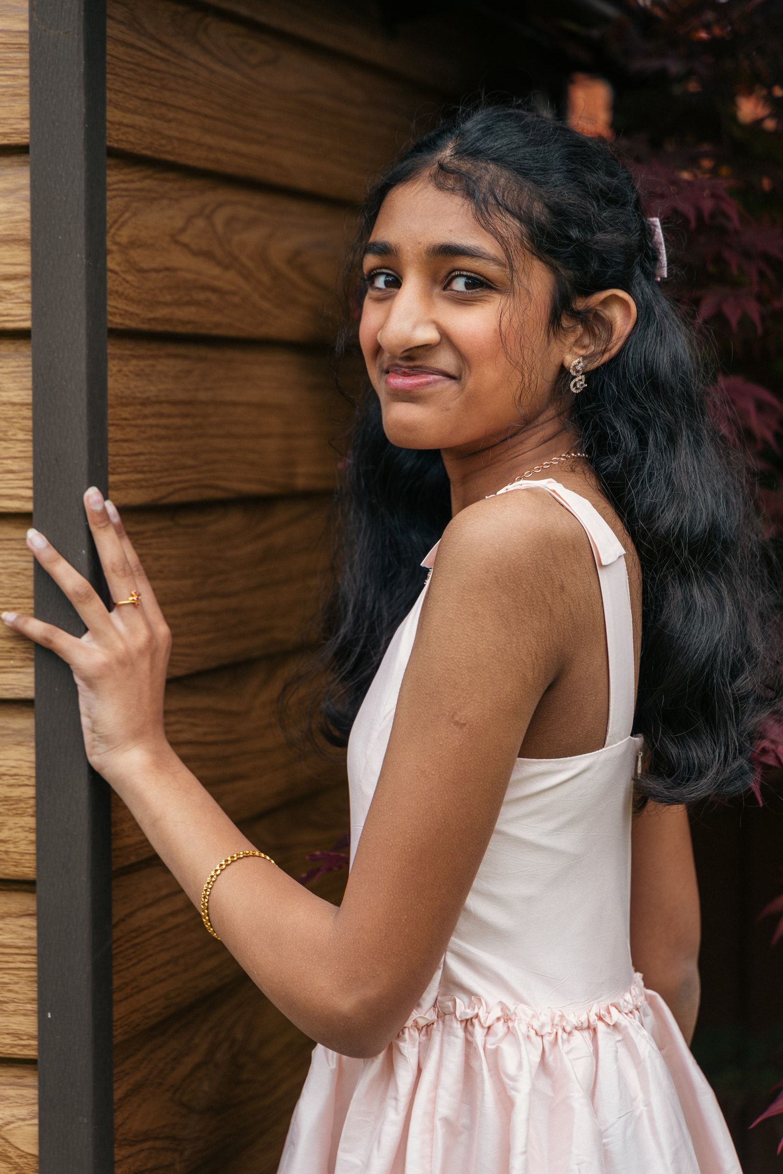 A young woman in a pale dress looking over her shoulder beside a wooden wall
