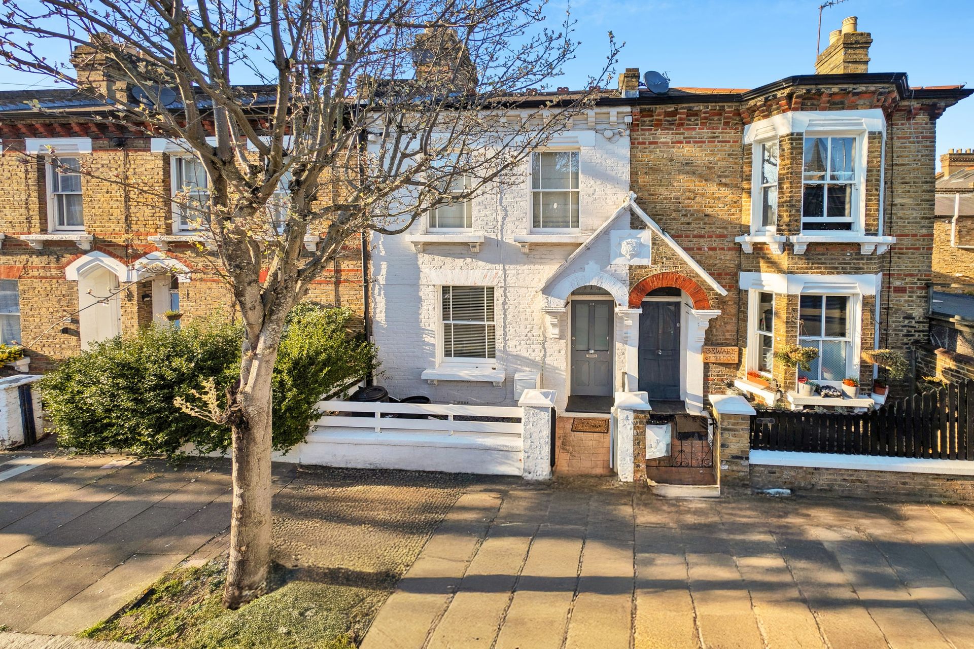 A sunlit terraced house with a white facade, front garden and tree beside the pavement